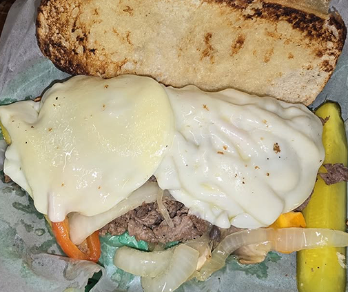 A steak sandwich on toasted bread with melted provolone, grilled onions and peppers, served with pickle spears