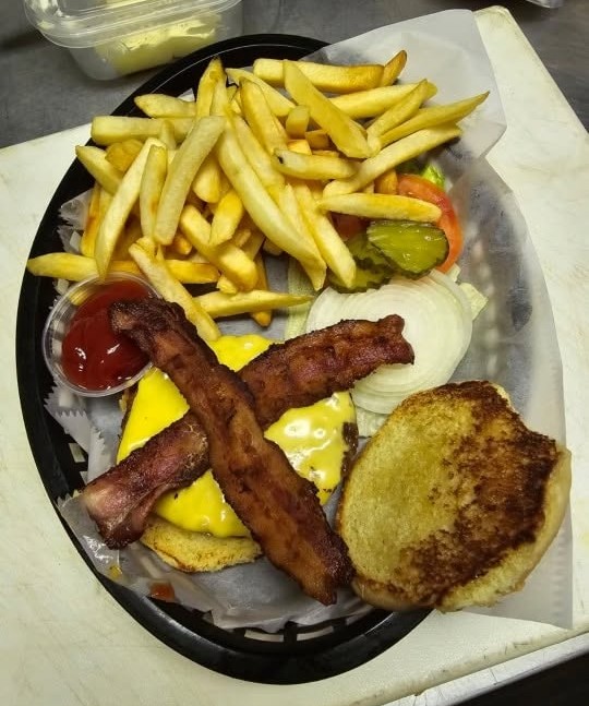 A bacon cheeseburger in a basket with french fries, lettuce, tomato, pickle, raw onion and a cup of ketchup