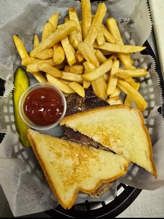 A hamburger patty served between slices of grilled Texas toast, in a basket with french fries, a pickle spear and a cup of ketchup